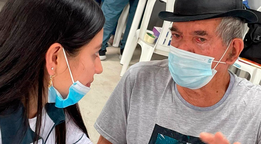 En la imagen, una persona joven conversa amablemente con una persona mayor que lleva sombrero y tapabocas. El encuentro refleja respeto y cuidado mutuo.