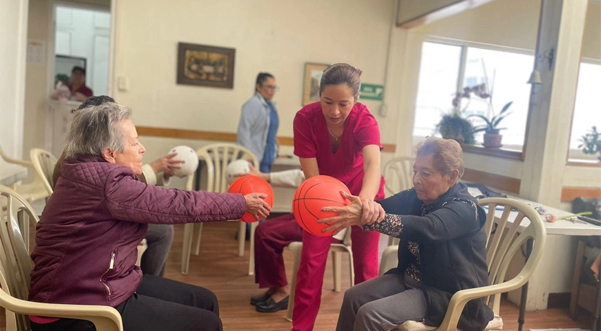Dos personas mayores realizan ejercicios de estiramiento utilizando balones de baloncesto, acompañadas y guiadas por una mujer enfermera. La escena transmite el compromiso con una vida activa a cualquier edad.